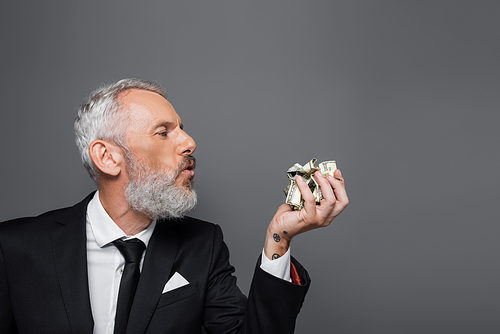 side view of bearded middle aged businessman blowing on crumpled dollar banknotes isolated on grey