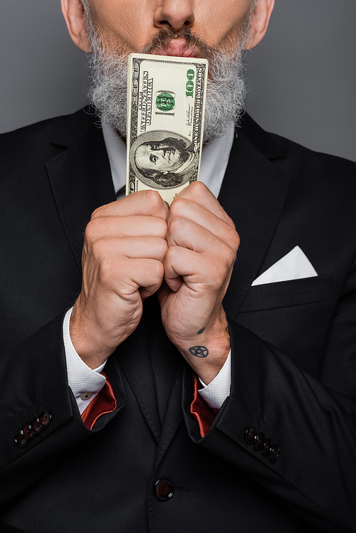 partial view of tattooed middle aged businessman kissing dollar banknotes isolated on grey