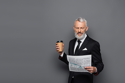 smiling middle aged businessman holding paper cup and reading newspaper on grey