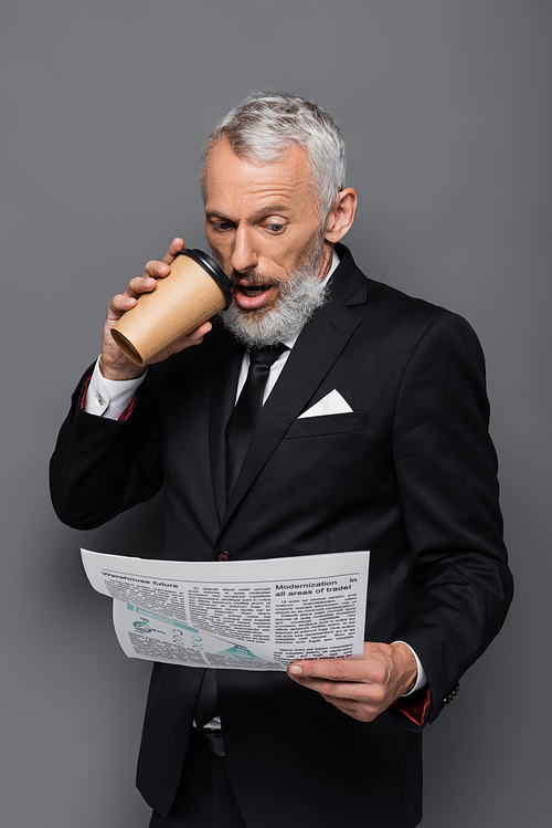 shocked middle aged businessman holding paper cup and reading newspaper on grey