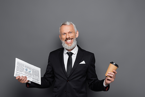 positive middle aged businessman holding paper cup and newspaper on grey