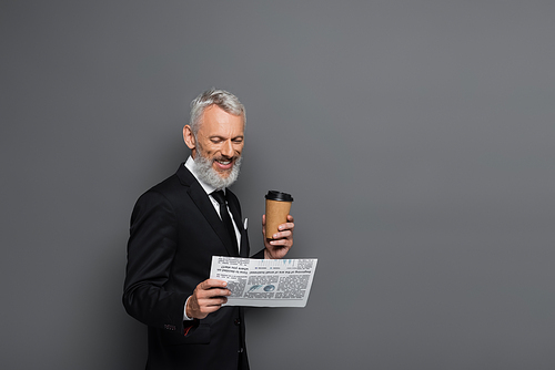 joyful middle aged businessman holding paper cup and reading newspaper on grey