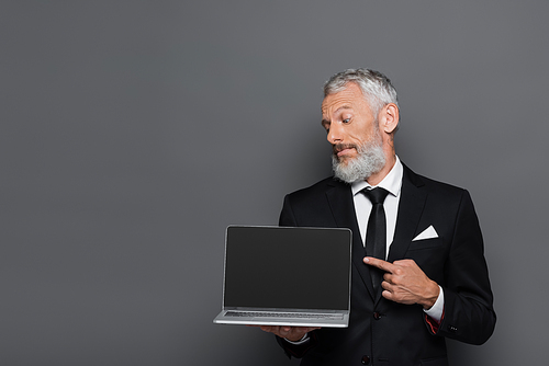 middle aged businessman in suit pointing at laptop with blank screen on grey