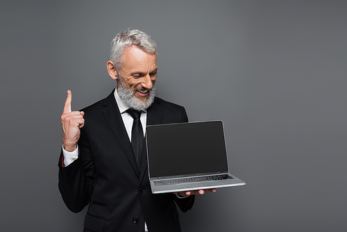 happy middle aged businessman in suit holding laptop with blank screen and pointing up isolated on grey