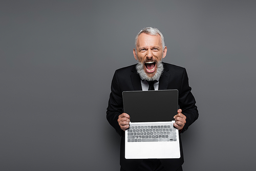 amazed middle aged businessman in suit holding laptop with blank screen on grey