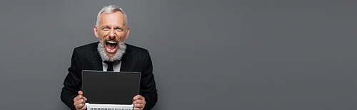 amazed middle aged businessman in suit holding laptop with blank screen on grey, banner