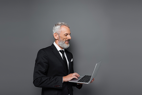 happy middle aged businessman in suit using laptop on grey