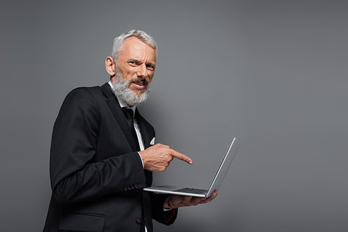 middle aged businessman in suit pointing with finger at laptop isolated on grey