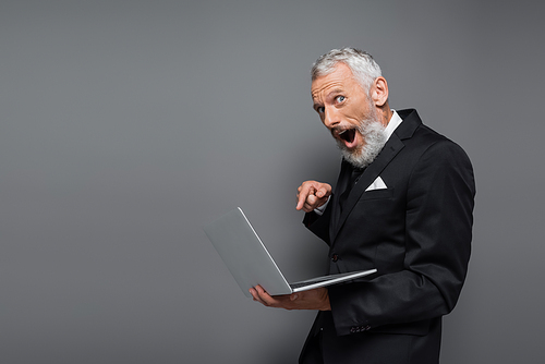 shocked middle aged businessman in suit pointing with finger at laptop on grey