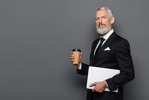 middle aged businessman in suit holding laptop and paper cup on grey