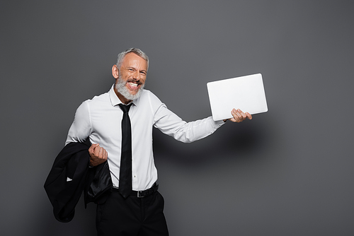 happy middle aged businessman holding laptop and blazer on grey