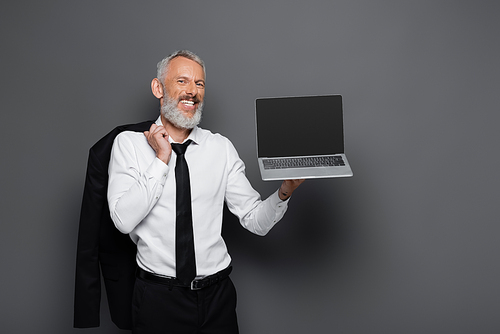 happy middle aged businessman in suit holding laptop with blank screen and blazer on grey