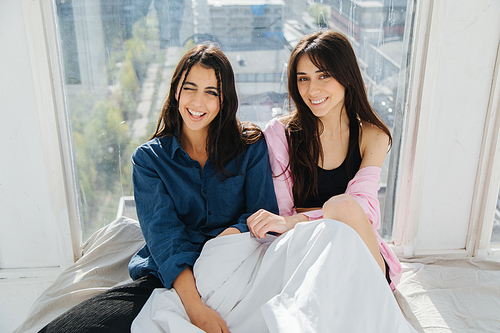 joyful armenian women smiling at camera while sitting near window on white bedding