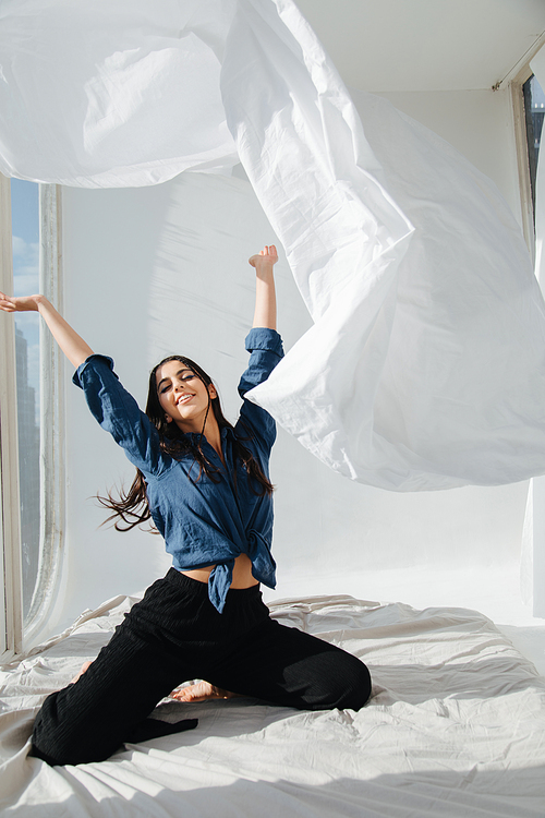 cheerful armenian woman throwing up white sheet in bedroom