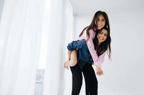 excited armenian women piggybacking happy friend near window at home