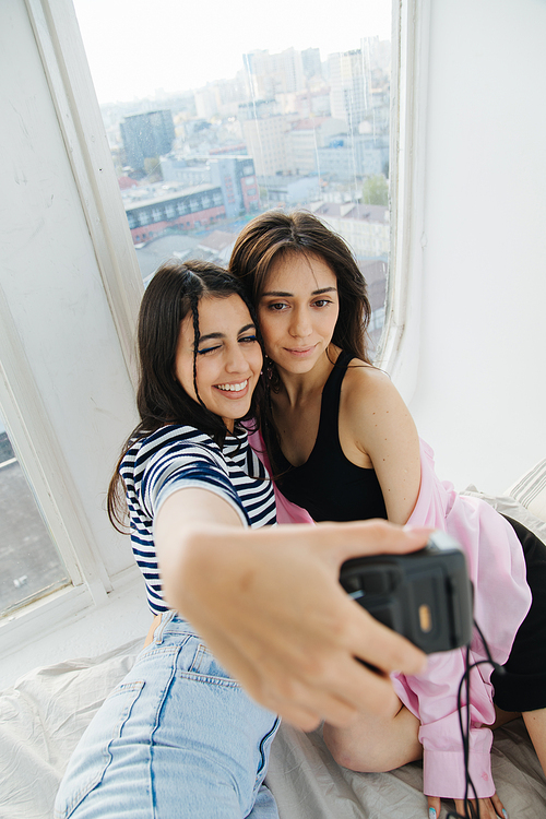 happy armenian woman taking selfie with friend on digital camera near window
