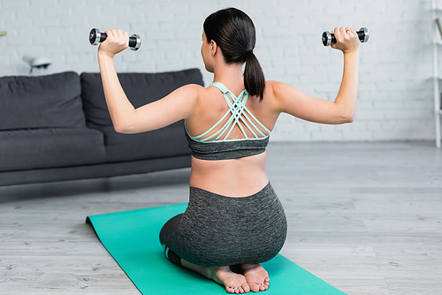 back view of woman in sportswear exercising with dumbbells while sitting on fitness mat