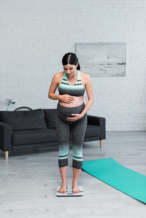 pregnant woman in sportswear hugging belly while measuring weight on floor scales