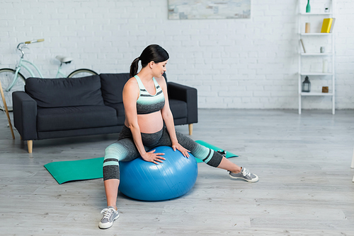 brunette pregnant woman in sportswear exercising on fitness ball in living room