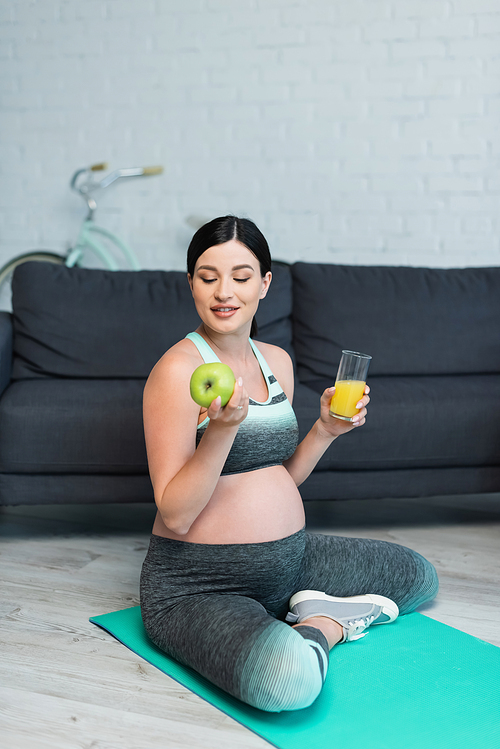 happy pregnant woman with apple and orange juice sitting with crossed legs on fitness mat