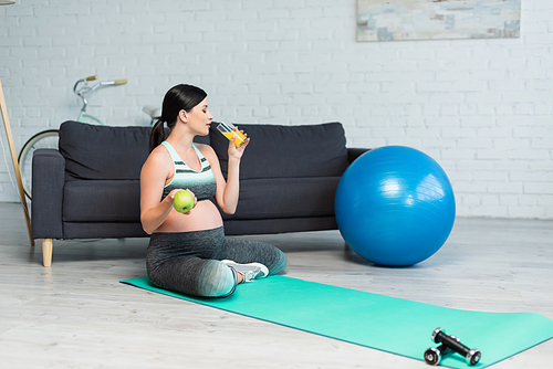 pregnant woman holding apple and drinking orange juice while sitting on fitness mat