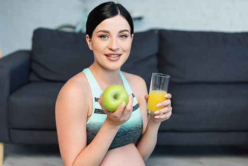young pregnant woman in sports bra smiling at camera while holding apple and orange juice