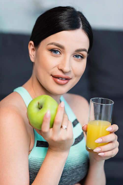 pretty, brunette woman holding ripe apple and fresh orange juice while smiling at camera