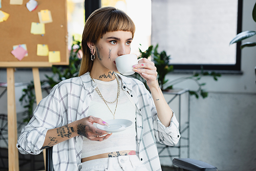 young tattooed businesswoman with piercing drinking coffee in office