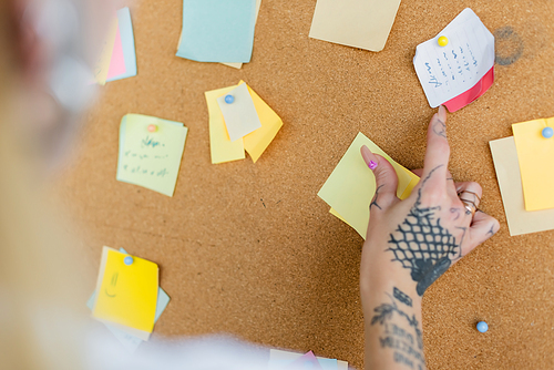 cropped view of blurred woman with tattoo near corkboard with sticky notes in office