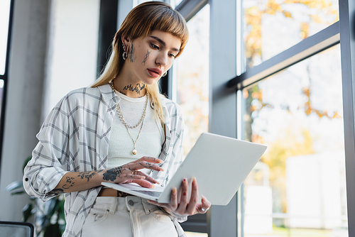 stylish tattooed businesswoman standing in office and typing on laptop