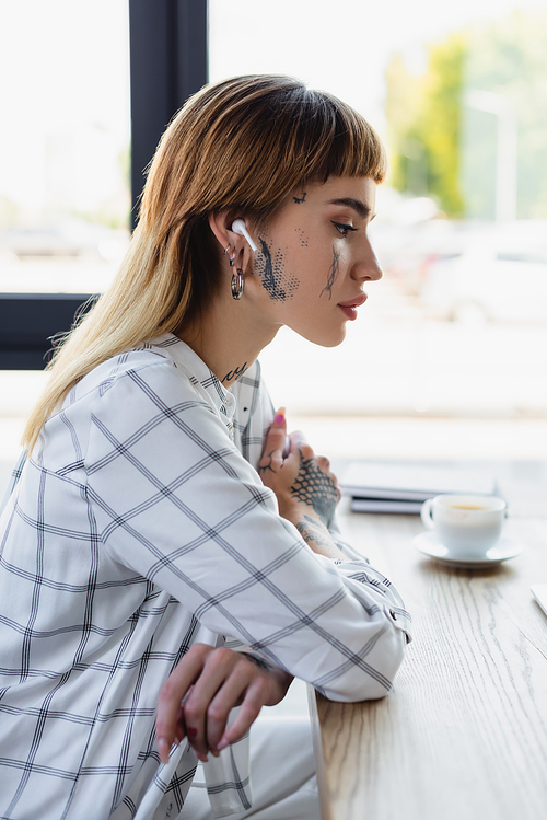 side view of tattooed woman in wireless earphone sitting at workplace with crossed arms
