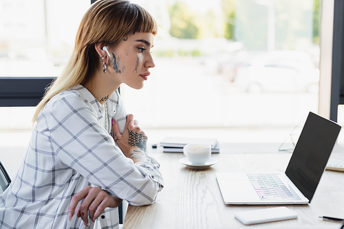 young tattooed businesswoman in earphone sitting with crossed arms near laptop with blank screen