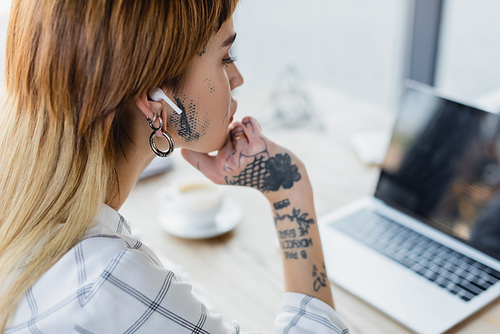 tattooed businesswoman in wireless earphone looking at blurred laptop in office