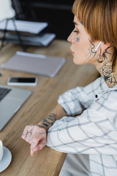 young tattooed woman in wireless earphone sitting at workplace near blurred gadgets