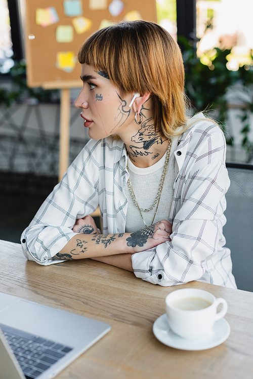 young woman with tattooed body sitting at work desk with crossed arms