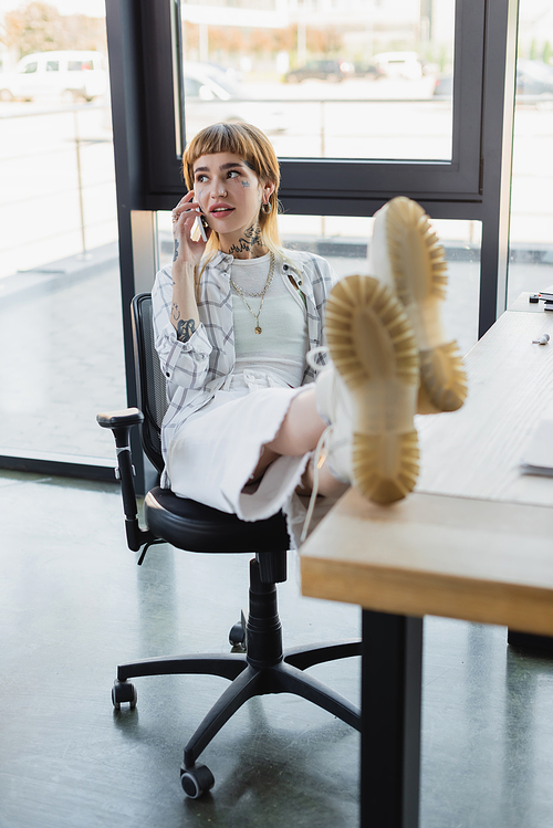 stylish tattooed woman sitting with legs on desk while talking on cellphone in office