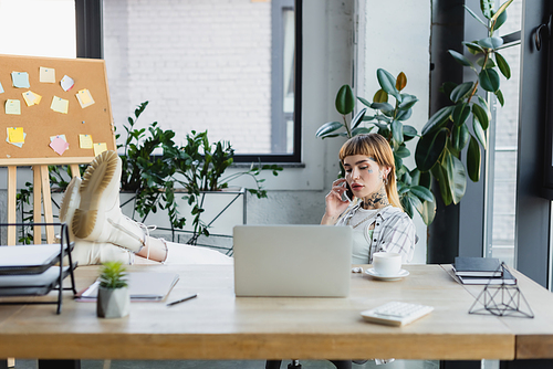 trendy businesswoman calling on cellphone while sitting with legs on desk near blurred laptop