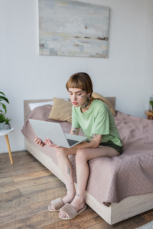 pretty woman with tattooed body working on laptop while sitting on bed at home