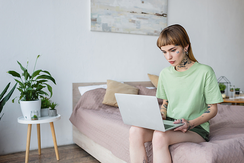 young woman with tattoo using laptop while sitting on bed at home