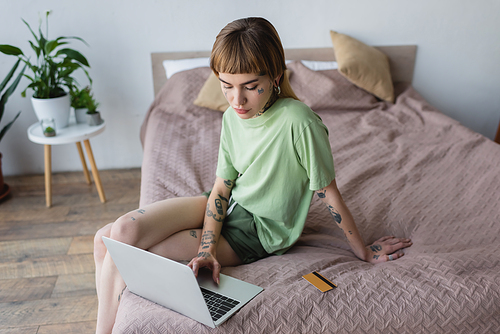young tattooed woman using laptop while sitting on bed near credit card