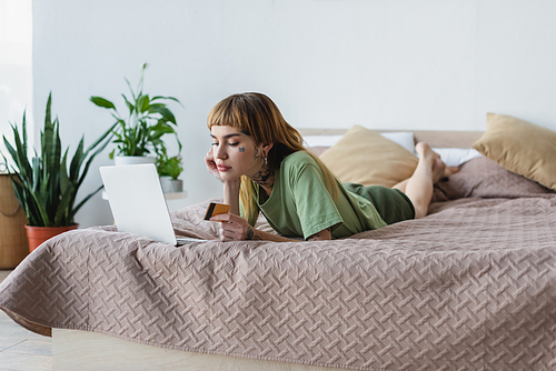 tattooed woman lying on bed with laptop and credit card near blurred plants in bedroom