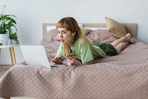 young woman with tattooed body lying on bed with credit card and laptop