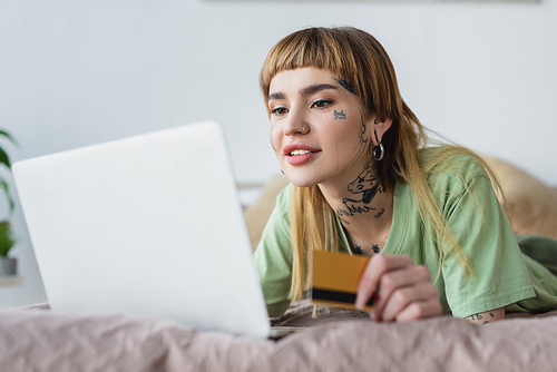 smiling tattooed woman holding credit card while lying on bed near blurred laptop