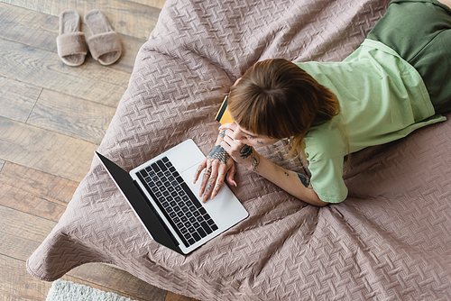 overhead view of tattooed woman using laptop while lying on bed with credit card