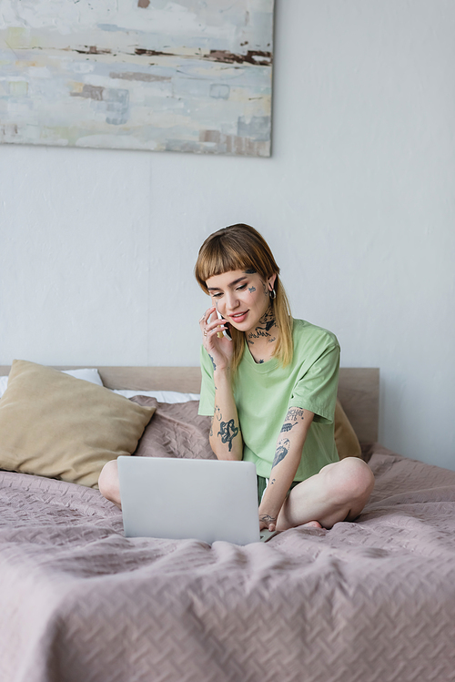 smiling tattooed woman working on laptop and talking on mobile phone on bed at home