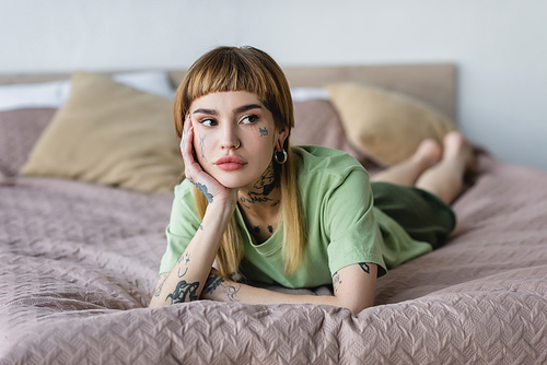 young woman with tattoo and piercing looking away while resting in bedroom