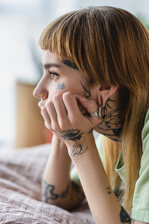side view of tattooed woman with piercing looking away on bed at home