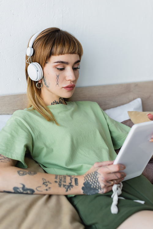 tattooed woman holding digital tablet while listening online lesson in headphones at home