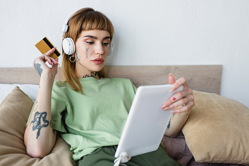 tattooed woman in headphones holding credit card while using digital tablet in bedroom