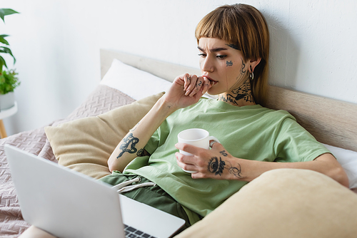 young worried woman with tattooed body watching film on blurred laptop